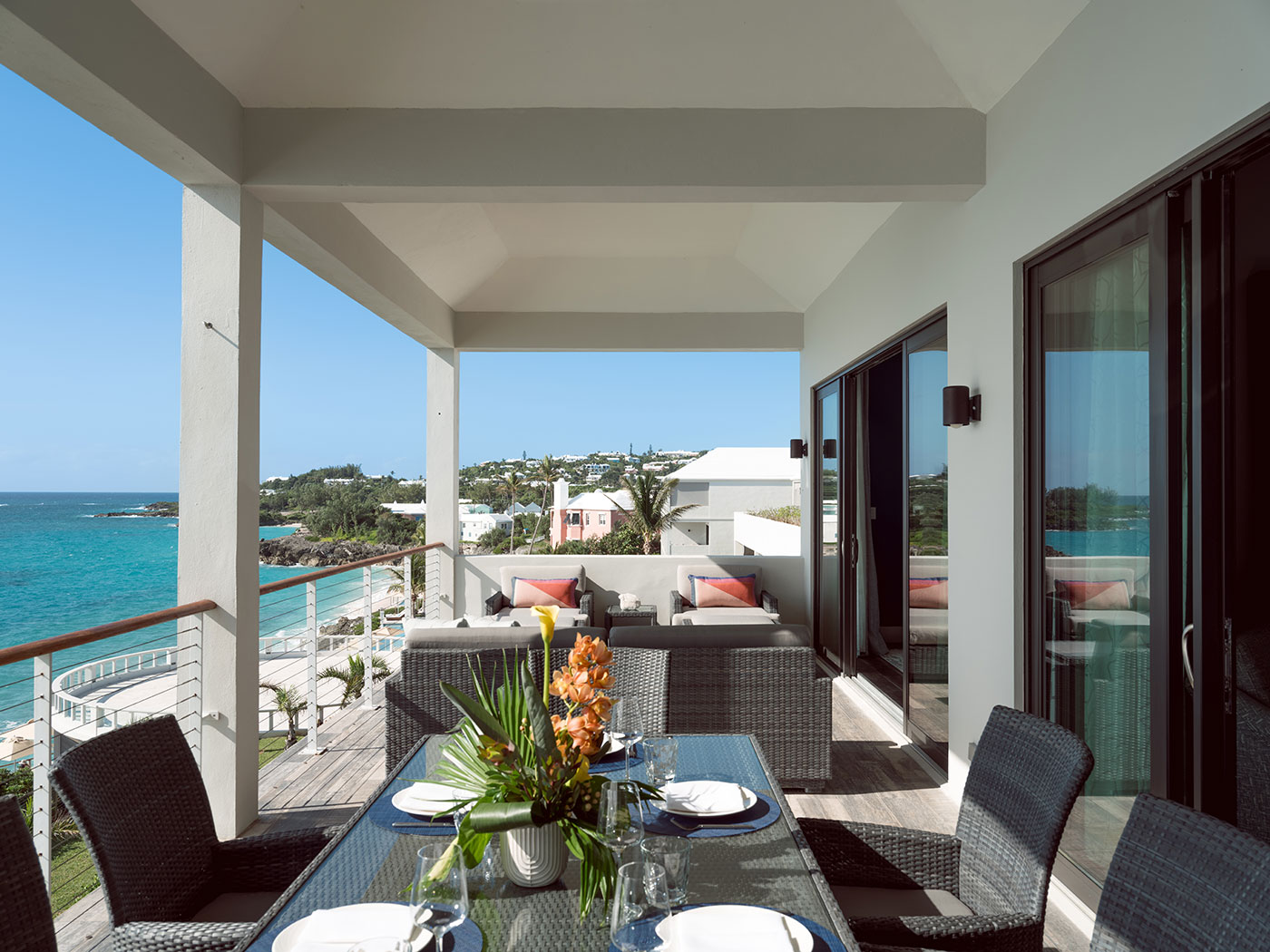 Modern covered balcony with outdoor dining table, wicker furniture, ocean view, and coastal buildings in the distance.