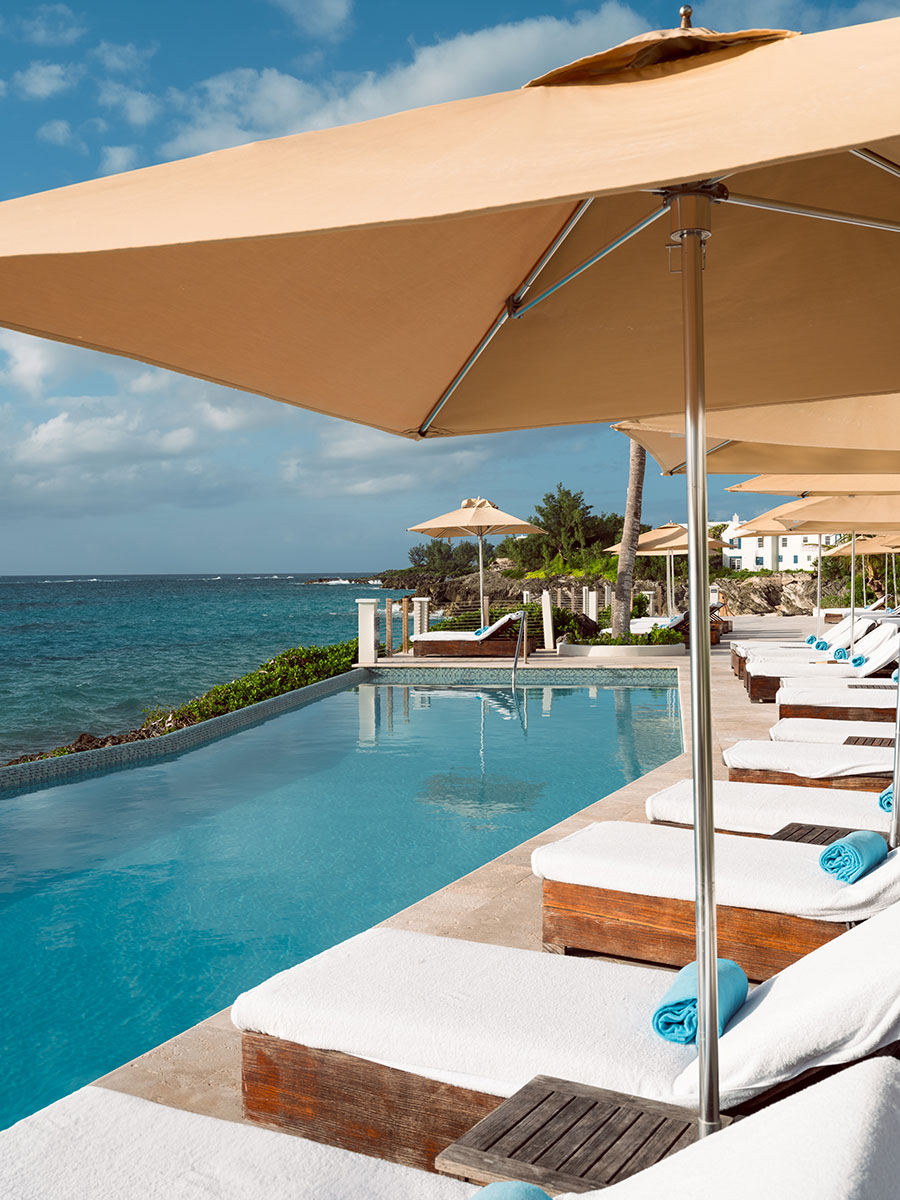 Poolside lounge chairs with towels under large umbrellas beside an infinity pool overlooking the ocean on a sunny day.