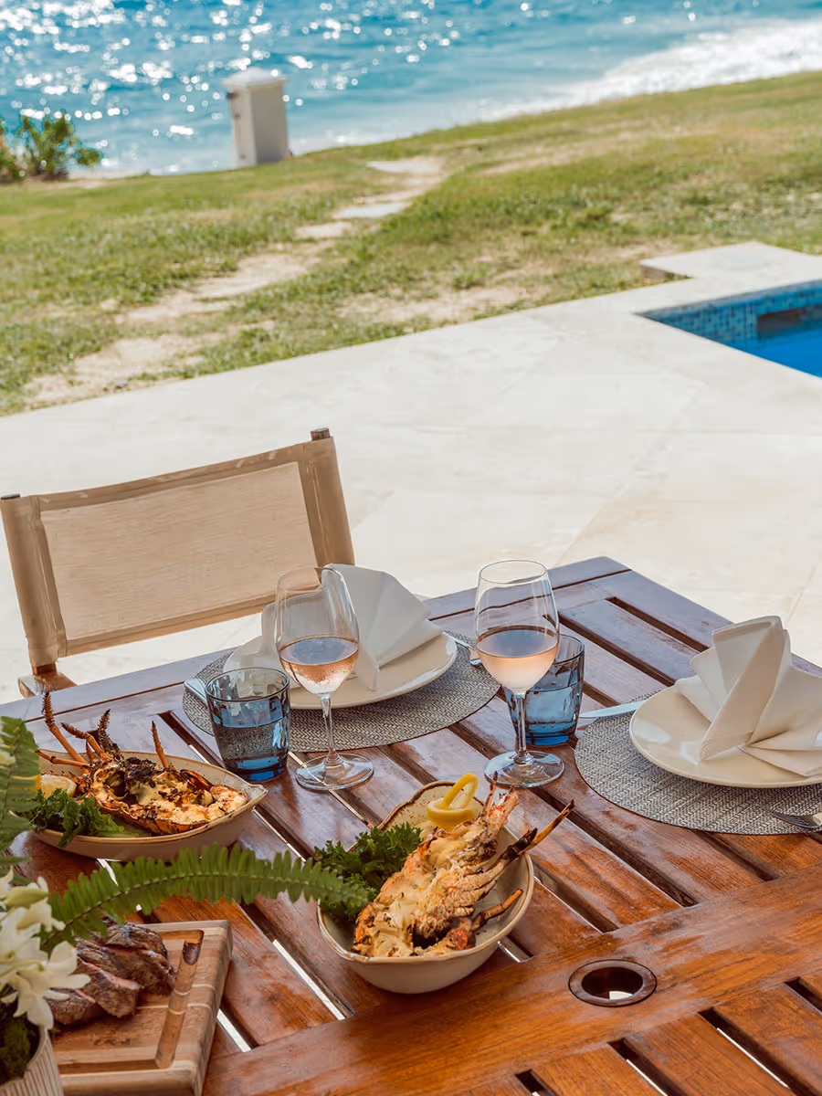 A wooden outdoor table is set with seafood dishes, wine glasses, and napkins, overlooking the ocean and a pool.