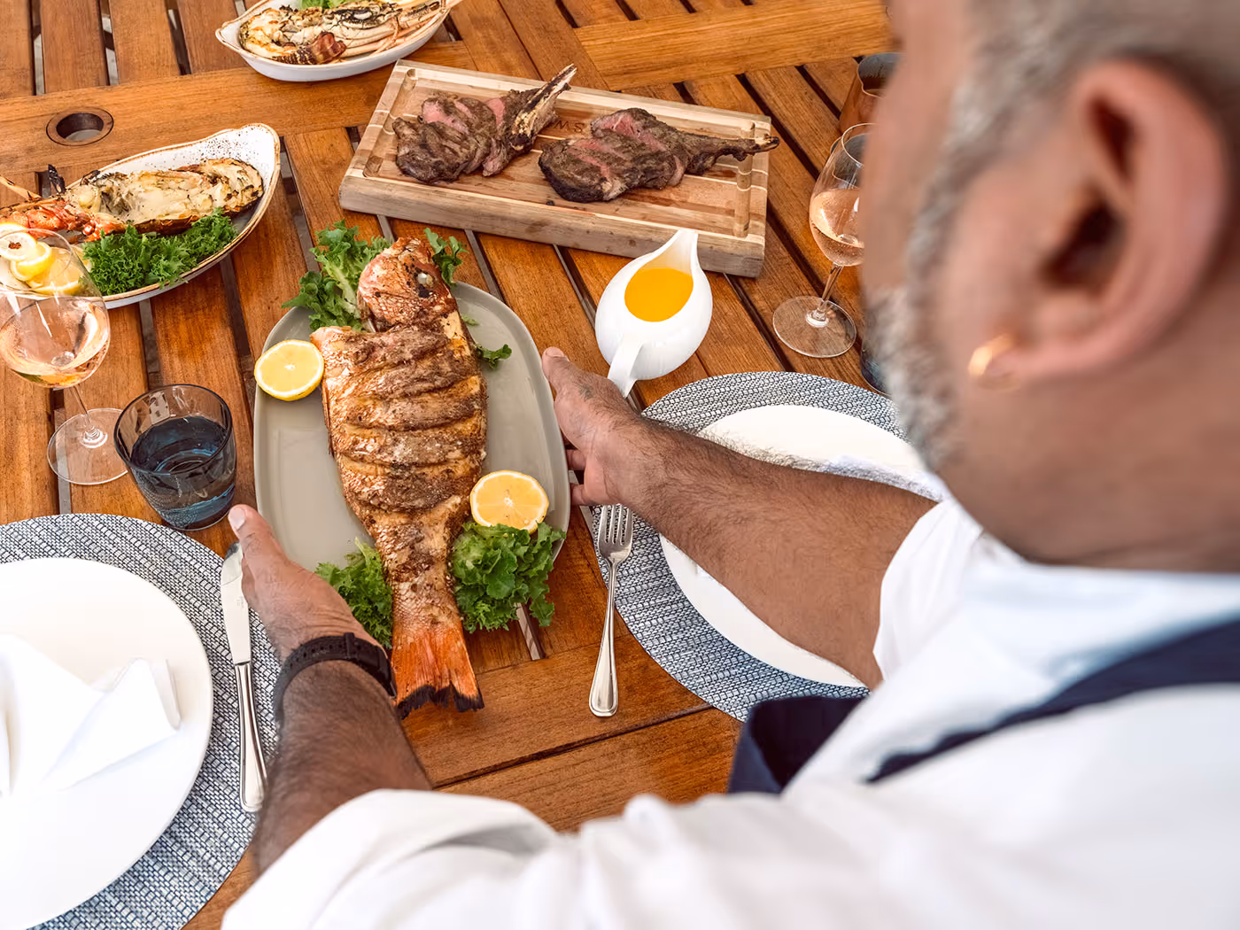 A person places a cooked whole fish on a table set with plates, steak, seafood, and wine glasses.