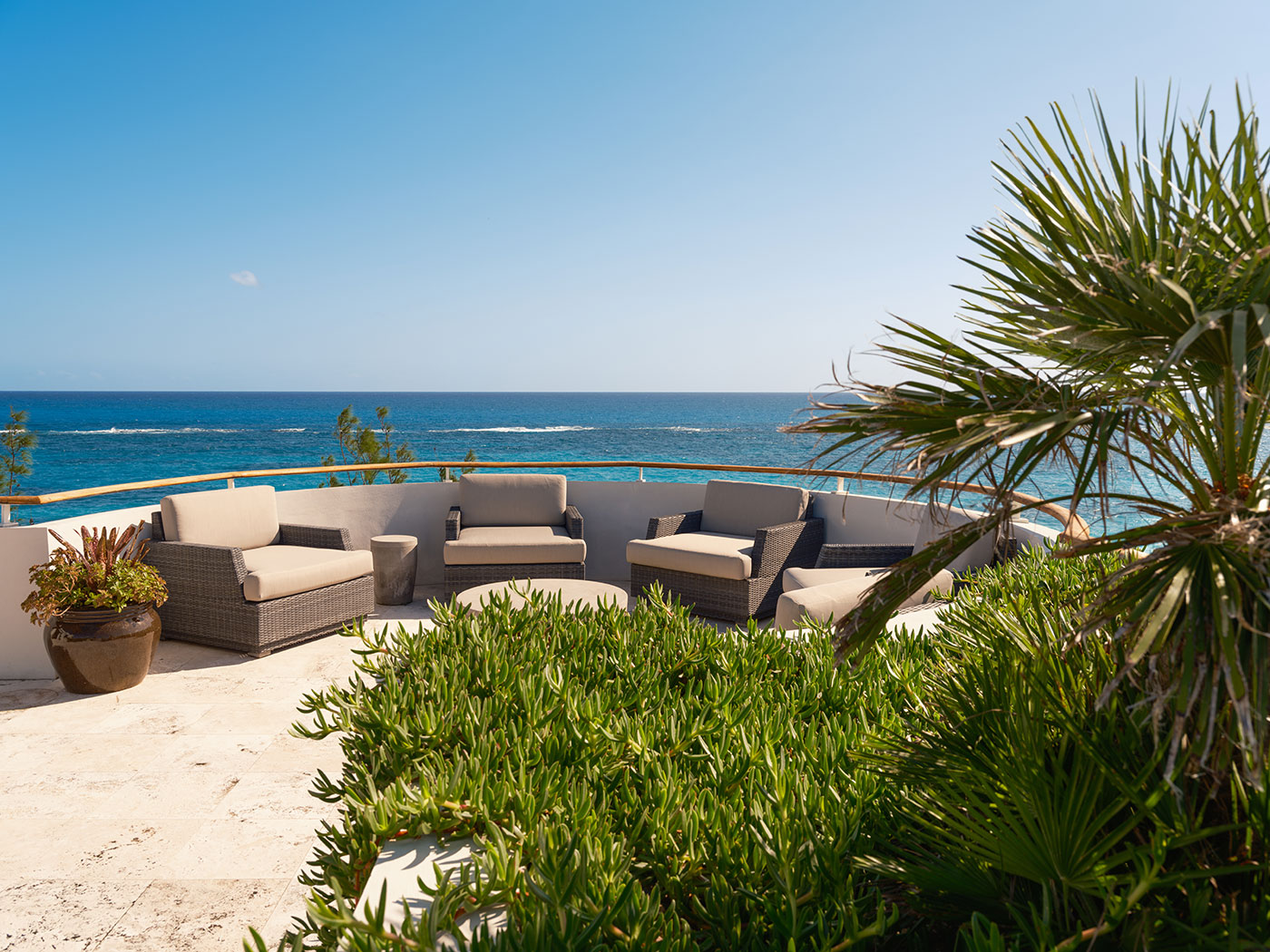 Outdoor patio with cushioned seating, tropical plants, and a view of the ocean under a clear blue sky.