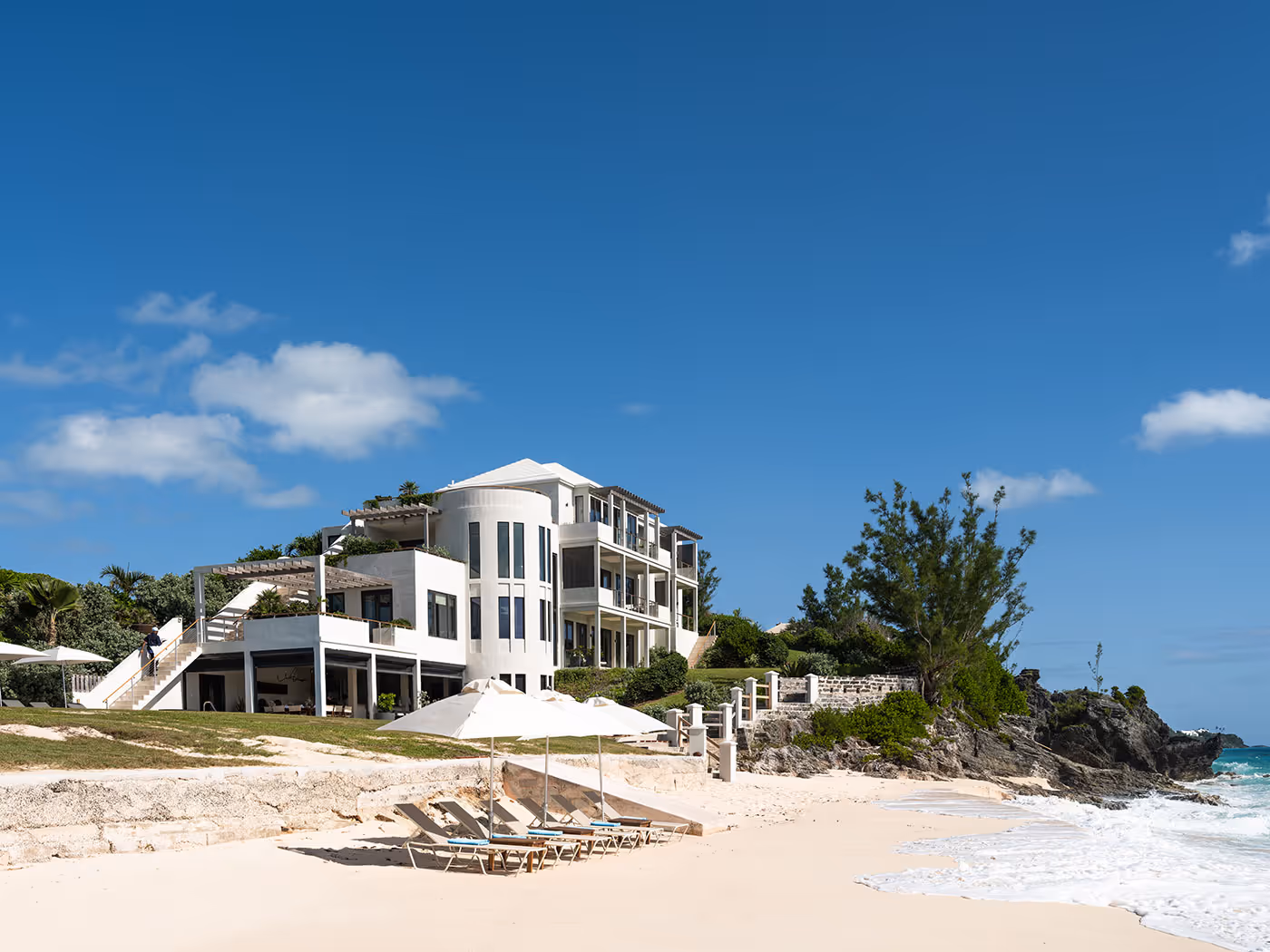 A modern white beachfront house with multiple balconies overlooks a sandy beach with lounge chairs and umbrellas.