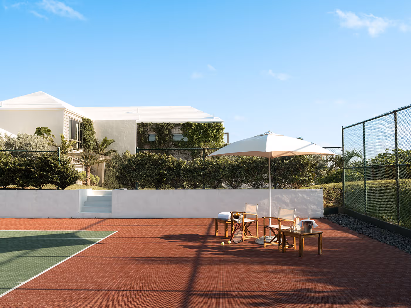 A tennis court with chairs and an umbrella on the side, surrounded by greenery and modern buildings in the background.