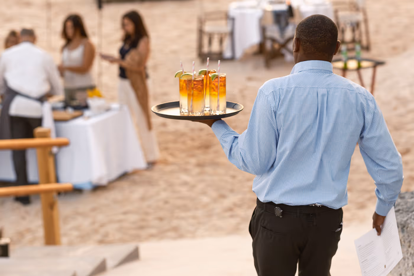 A server carries a tray of iced drinks with straws at an outdoor event with people and tables in the background.