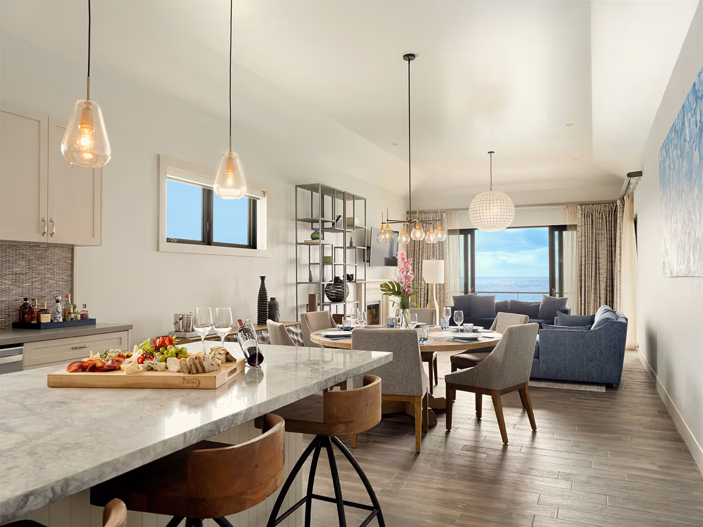 Modern open-concept kitchen and living area with marble island, dining table, and large windows overlooking water views.