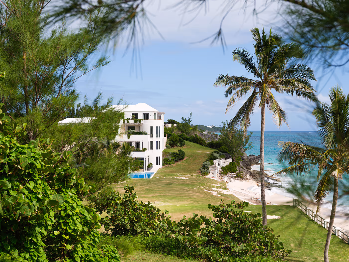 White three-story beachfront building surrounded by greenery, palm trees, and overlooking the ocean under a blue sky.