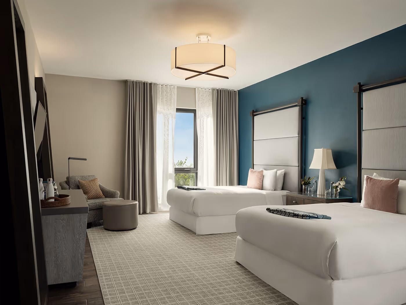 Modern hotel room with two double beds, a teal accent wall, seating area, large window, and neutral decor.