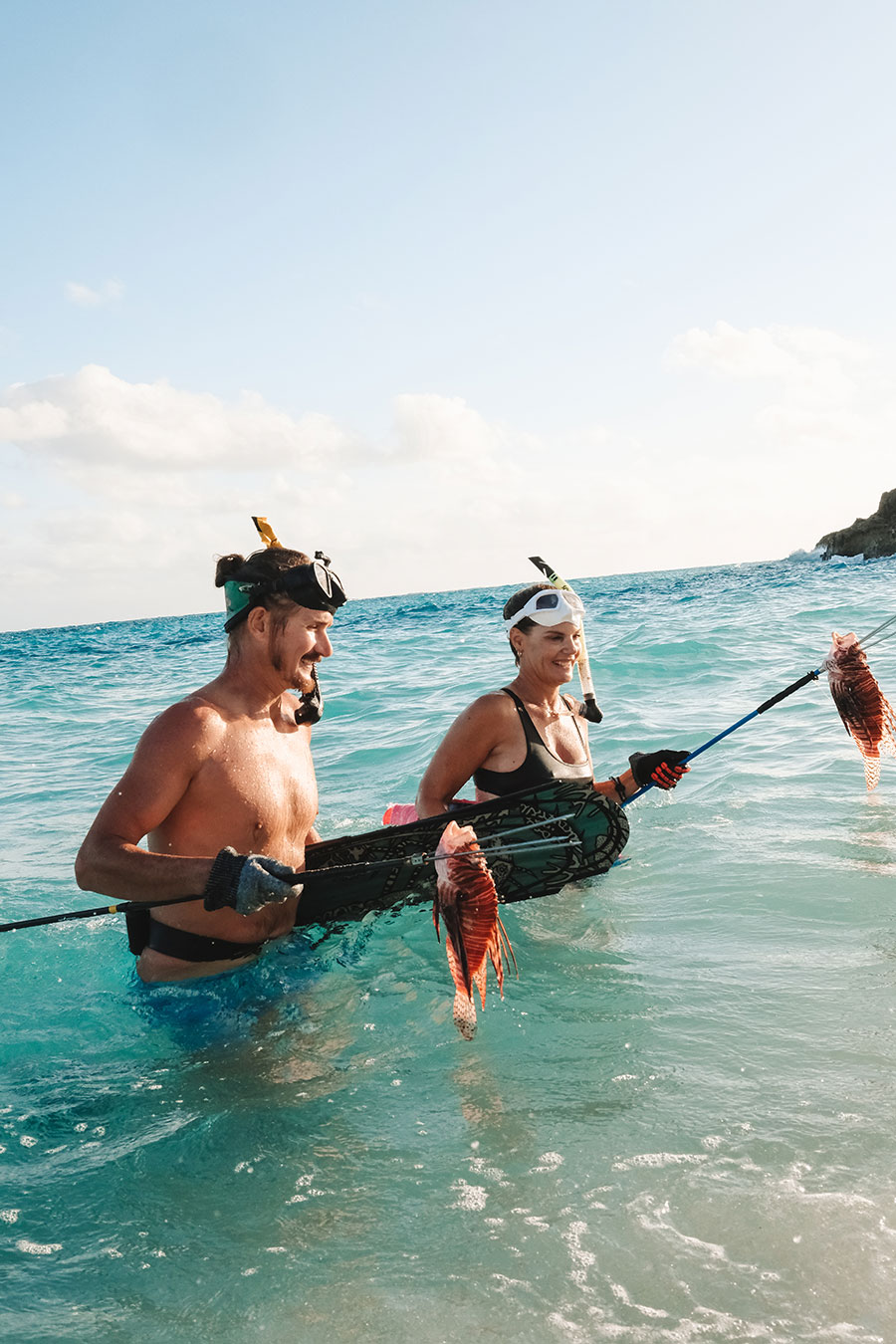 Two people in snorkel gear wade in ocean water, each holding a spear with a caught lionfish.