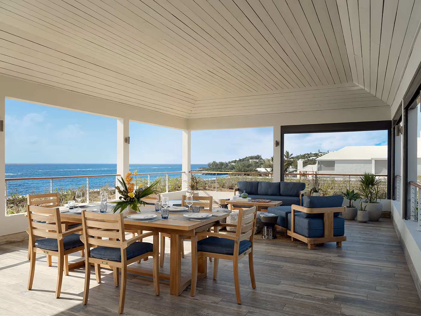 Open-air patio with wooden dining set and lounge area, overlooking the ocean and a modern coastal landscape.