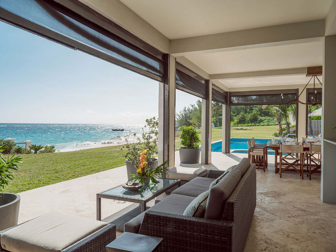 Modern covered patio with wicker furniture overlooks grassy lawn, pool, and ocean under clear blue sky.