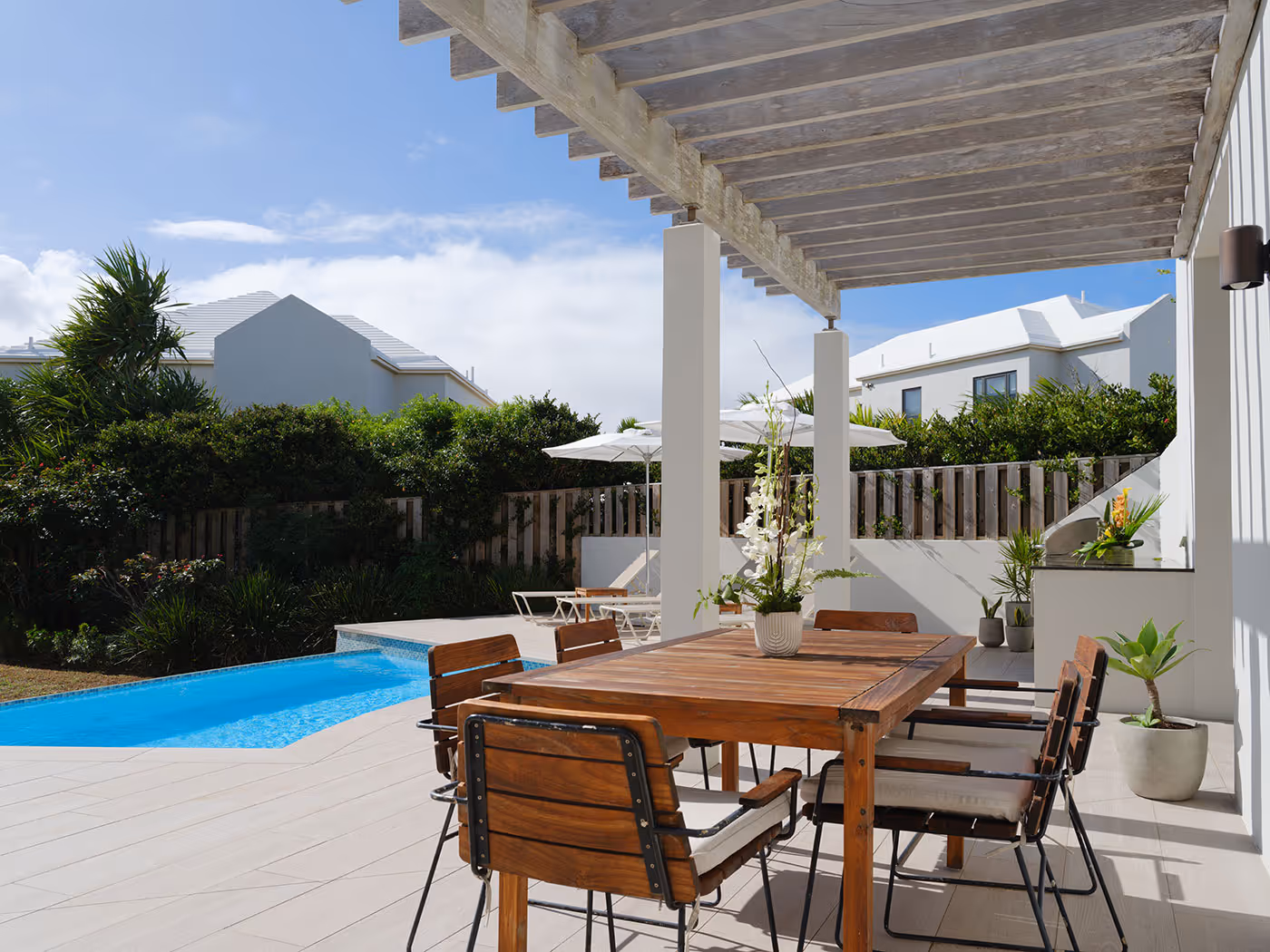 Wooden outdoor dining set under a pergola next to a pool in a modern backyard with plants and white houses in the background.