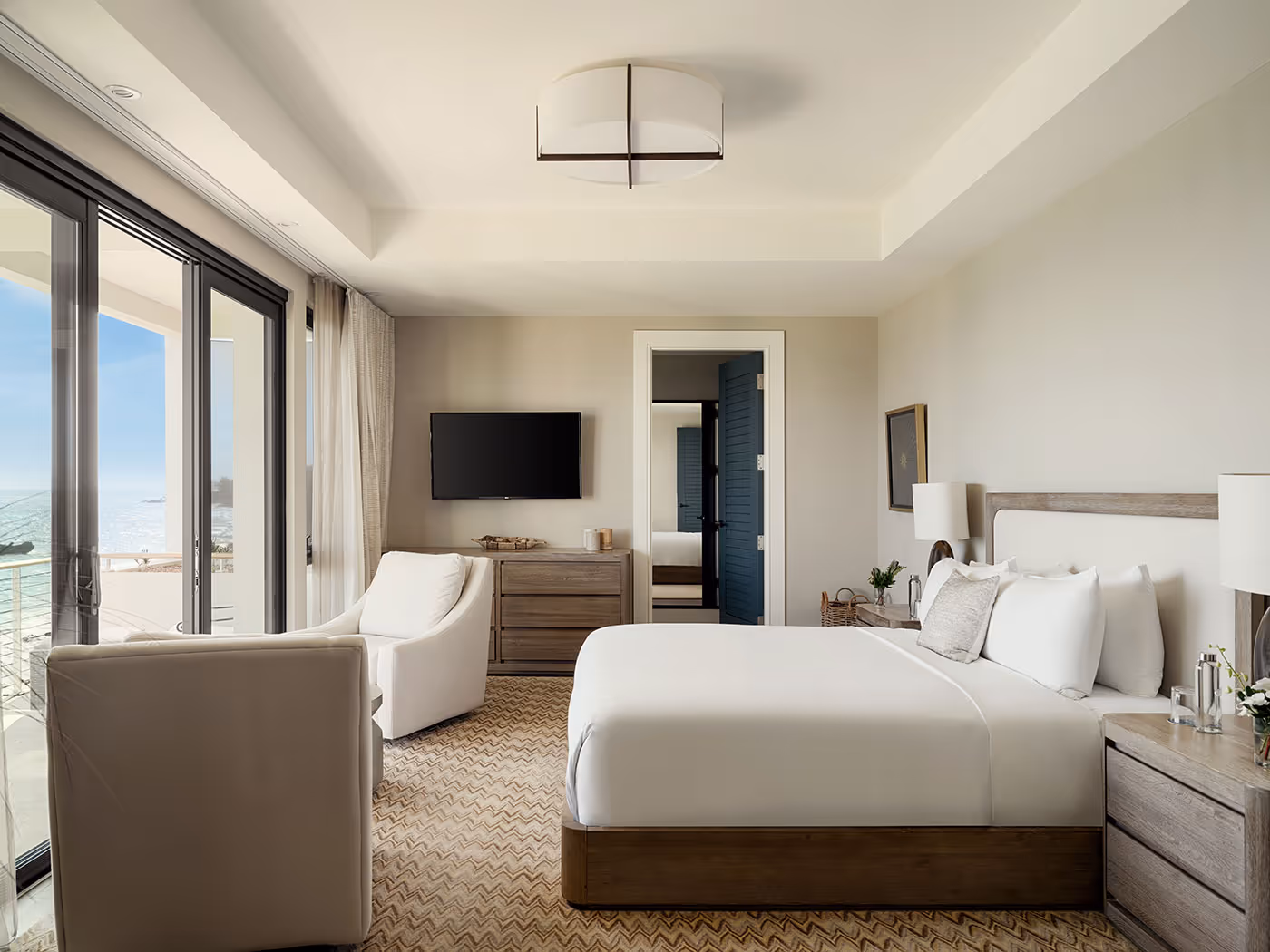 Modern hotel room with a large bed, armchairs, TV, dresser, and balcony with ocean view.