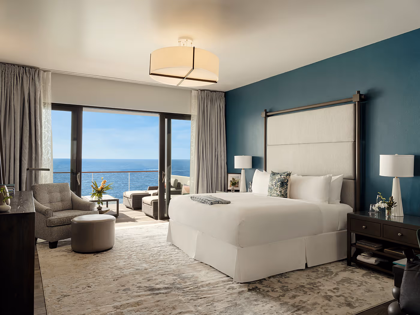 Modern hotel room with large bed, seating area, and ocean view through sliding glass doors opening to a balcony.