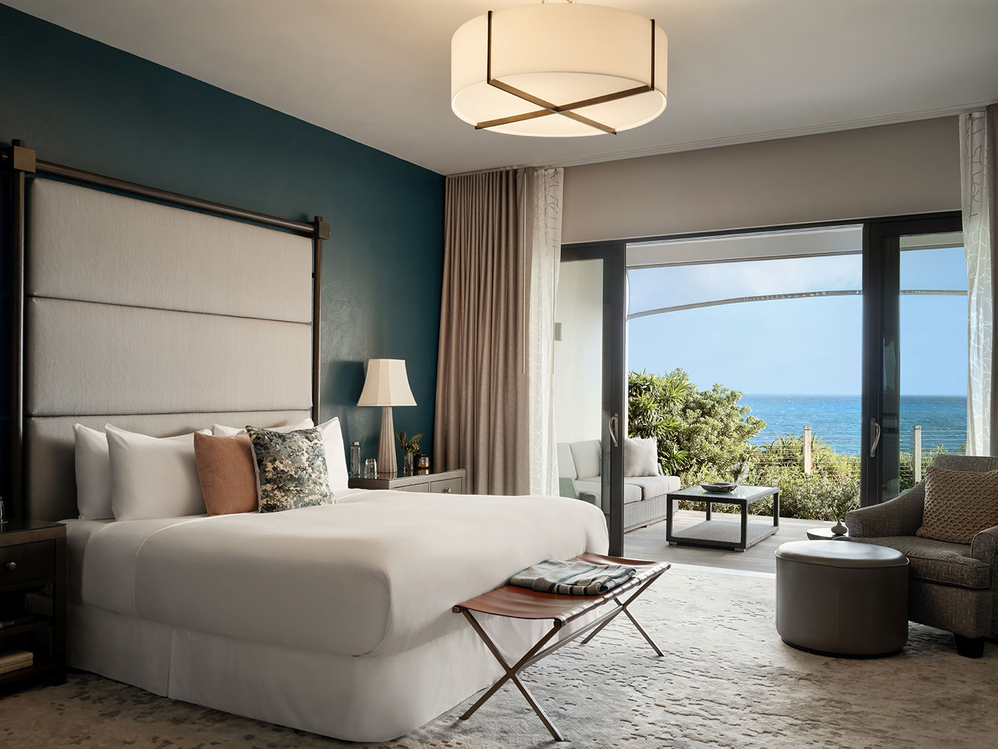 Modern hotel bedroom with large bed, seating area, and glass doors opening to a patio with an ocean view.