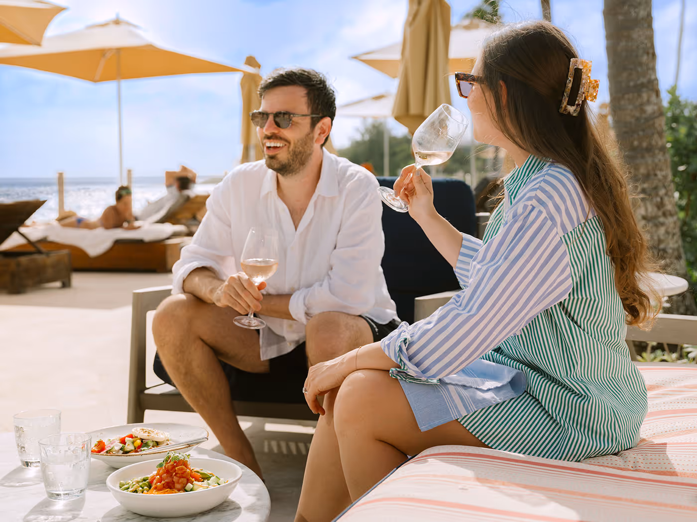 Two people sit outdoors on a lounge, eating pasta and drinking wine, with sun umbrellas and beach chairs in the background.