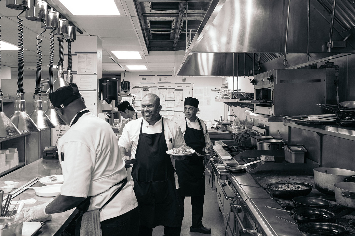 Chefs work together in a busy restaurant kitchen, preparing and plating food near stoves and counters.