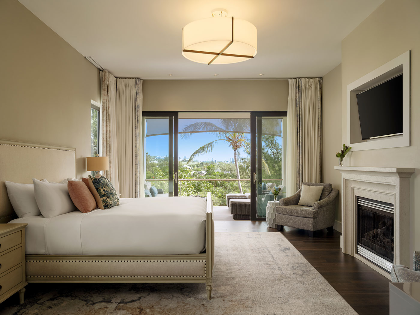 Modern bedroom with a large bed, fireplace, TV, seating area, and glass doors opening to a balcony with a palm tree view.