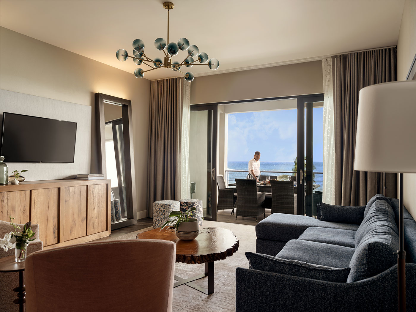 Modern hotel suite with a blue sofa and balcony view; a man stands outside overlooking the ocean.
