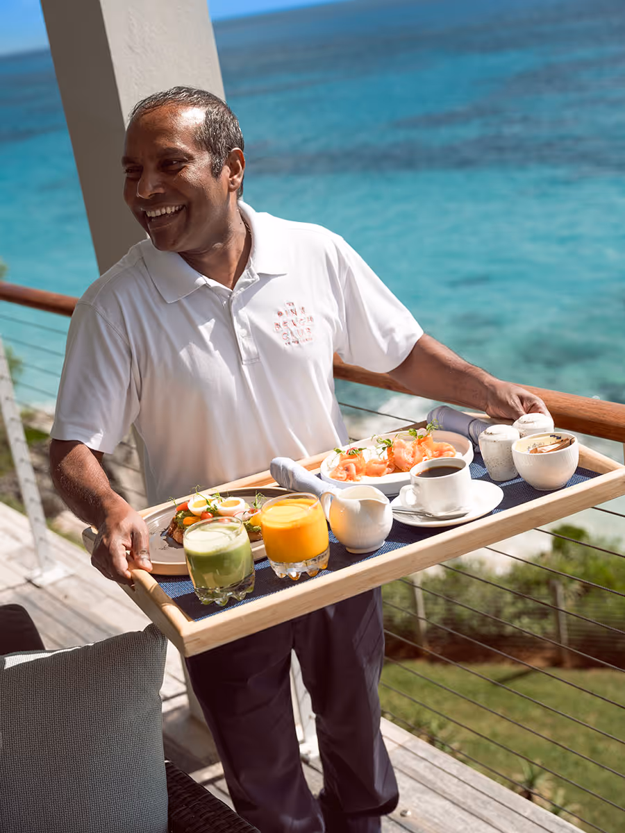 A server carrying a tray with breakfast plates, juice, and coffee on a balcony overlooking the ocean.