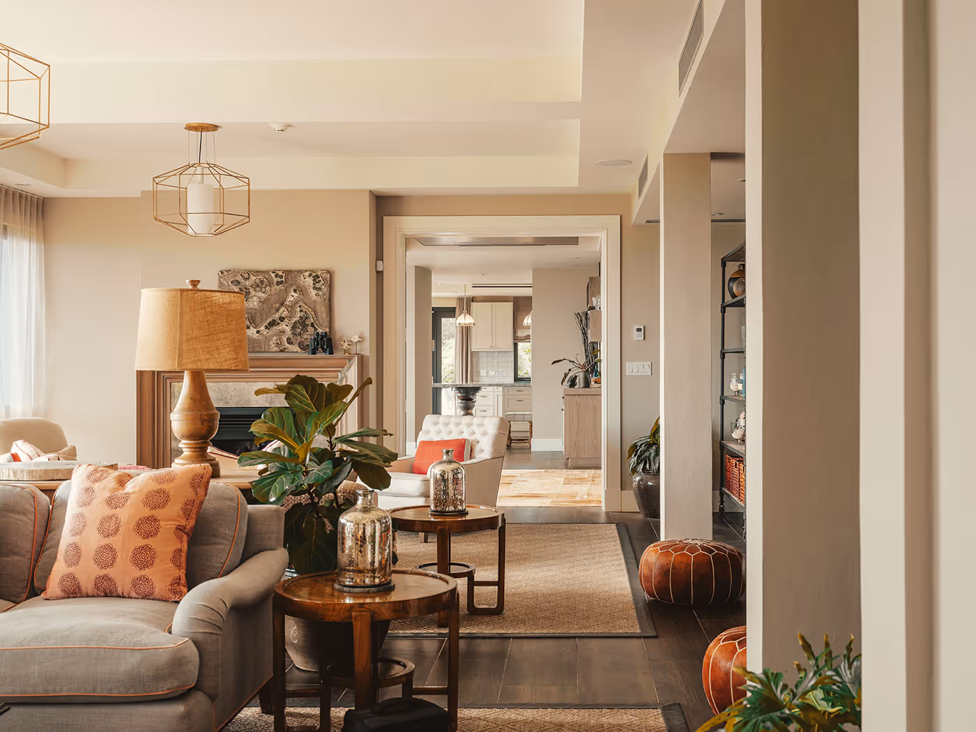 Modern living room with neutral tones, patterned pillows, plants, and a view into a bright kitchen area in the background.