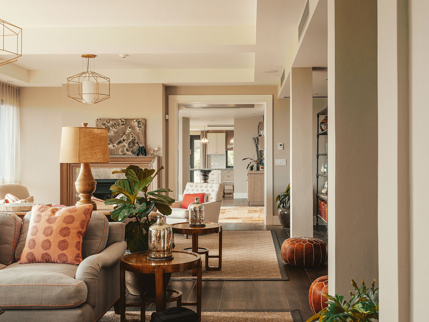 Modern living room with neutral tones, patterned pillows, plants, and a view into a bright kitchen area in the background.
