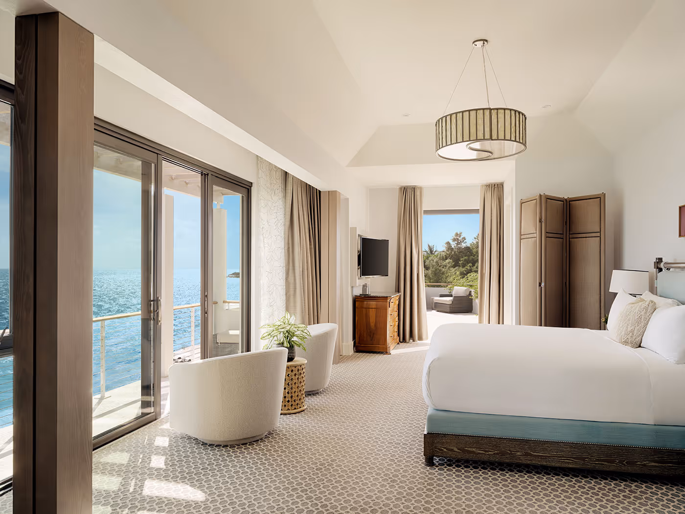 Spacious hotel room with large bed, two chairs, a TV, and balcony overlooking the ocean under bright natural light.