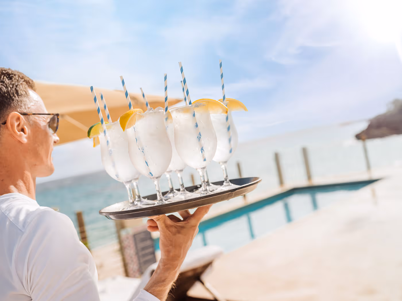 Server carrying a tray of six cocktails with striped straws and lemon wedges by a pool with an ocean view.