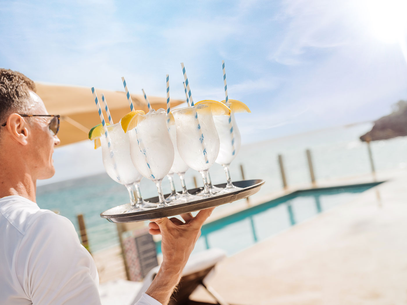 Server carrying a tray of six cocktails with striped straws and lemon wedges by a pool with an ocean view.