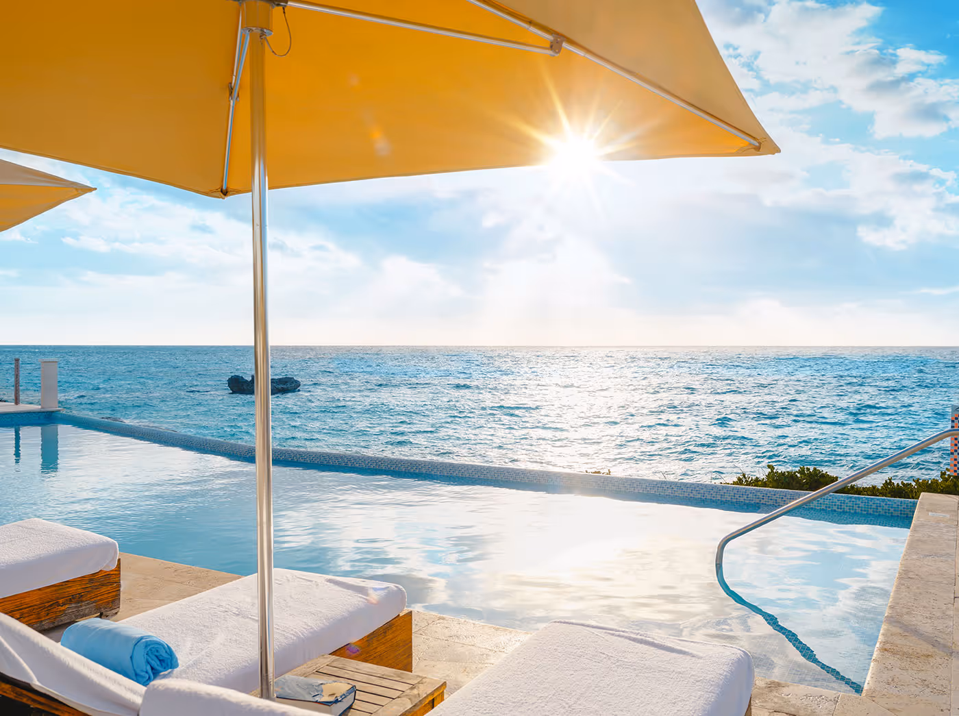 Infinity pool with sun loungers under yellow umbrellas overlooking the ocean at sunrise or sunset.