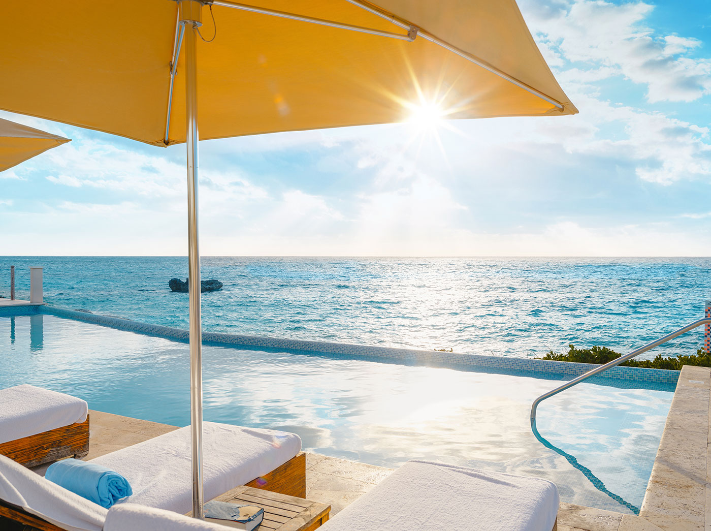 Infinity pool with sun loungers under yellow umbrellas overlooking the ocean at sunrise or sunset.