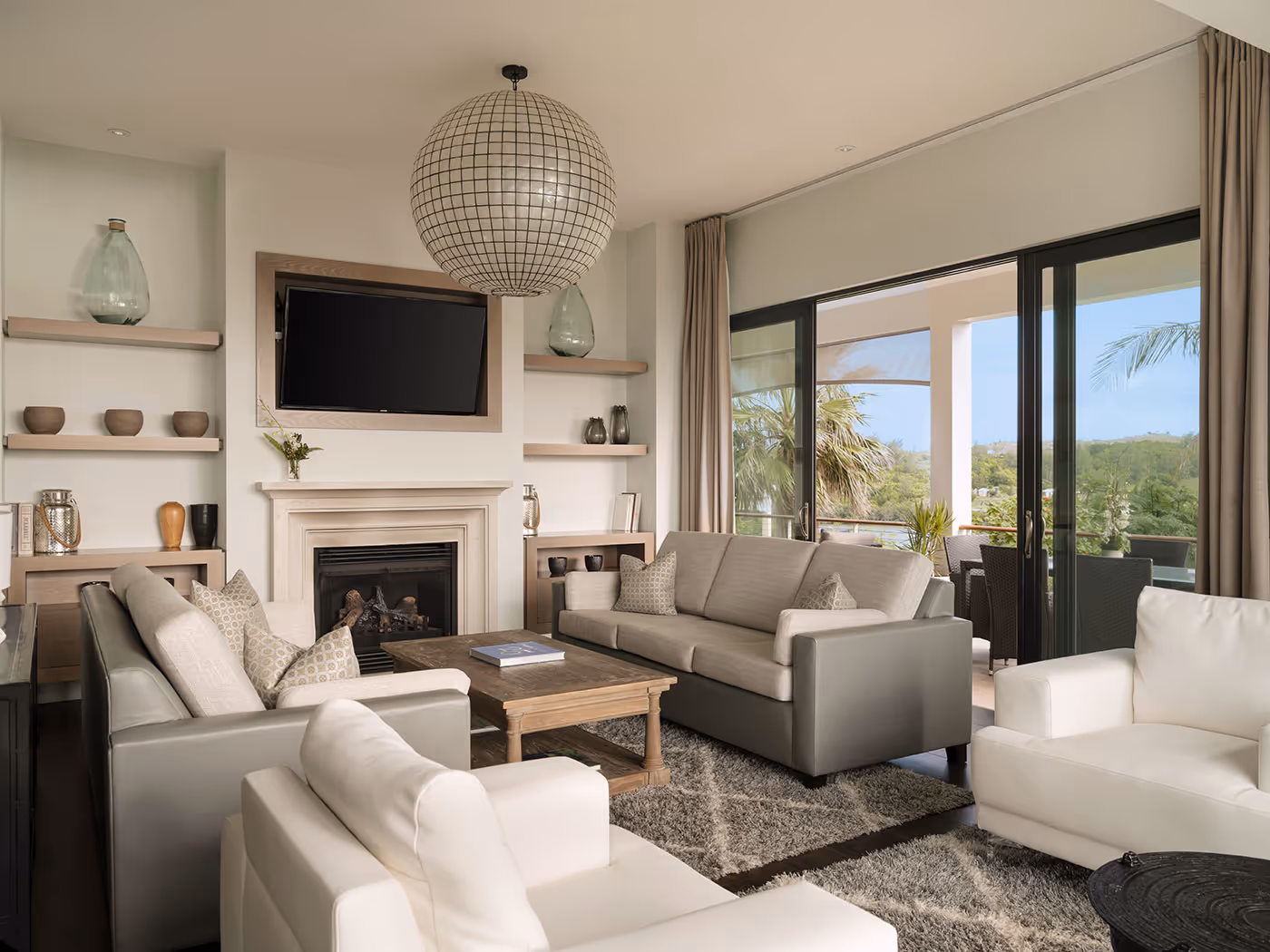 Modern living room with neutral furnishings, a fireplace, wall-mounted TV, and large windows overlooking a patio and greenery.