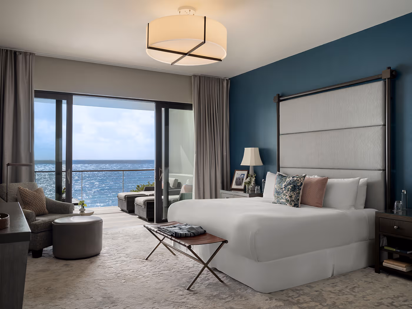 Modern hotel bedroom with a large bed, ocean view, and balcony seating area visible through sliding glass doors.