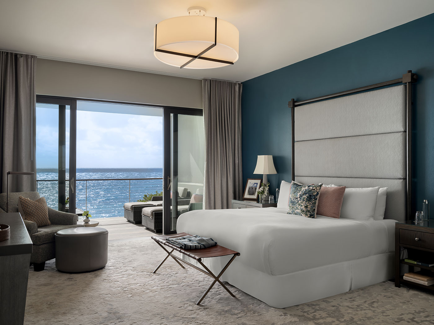 Modern hotel bedroom with a large bed, ocean view, and balcony seating area visible through sliding glass doors.