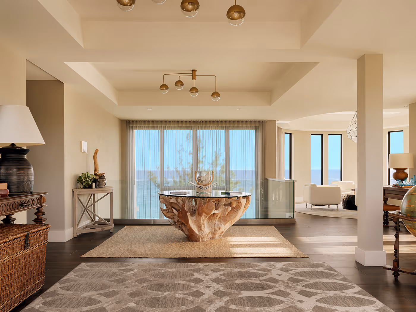 Bright, modern living room with large windows, unique wood table, neutral tones, and ocean view in the background.
