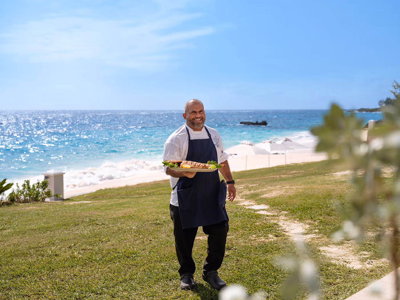 A chef in an apron stands on grass by the beach, holding a tray of food with the ocean and umbrellas in the background.