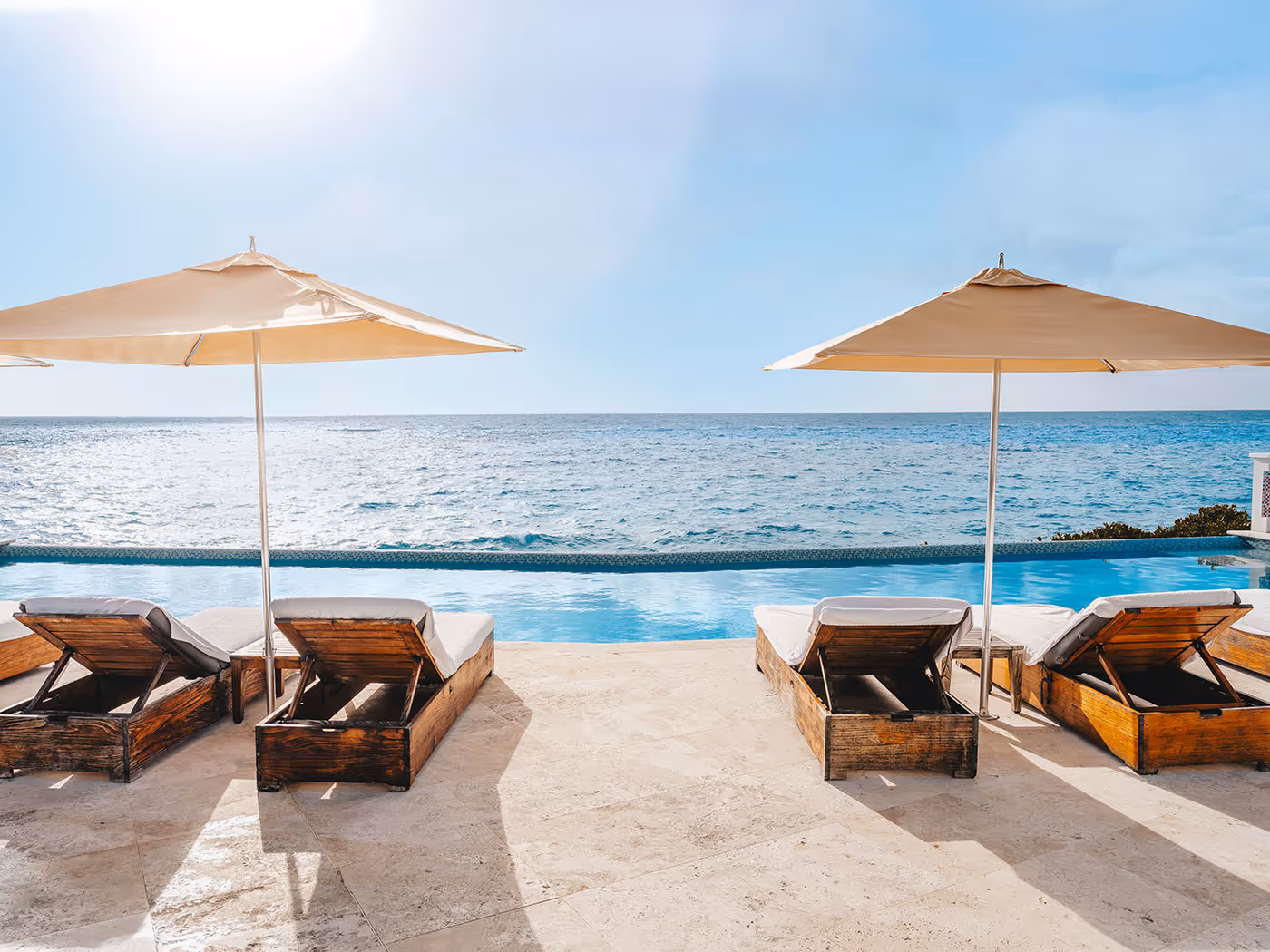 Four wooden lounge chairs under umbrellas face an infinity pool and ocean on a sunny day.