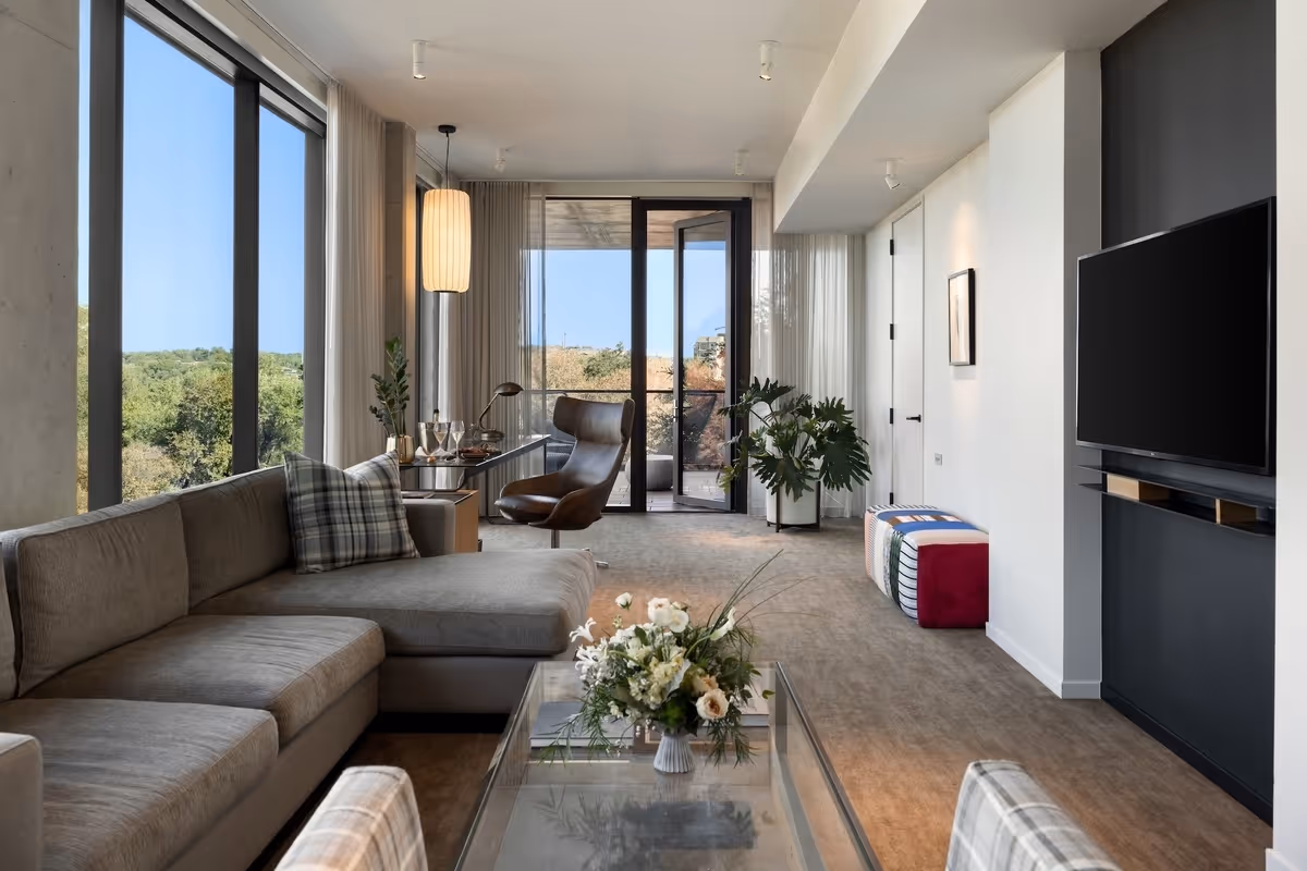 Modern living room with large windows, gray sectional sofa, armchair, wall-mounted TV, and glass coffee table with flowers.