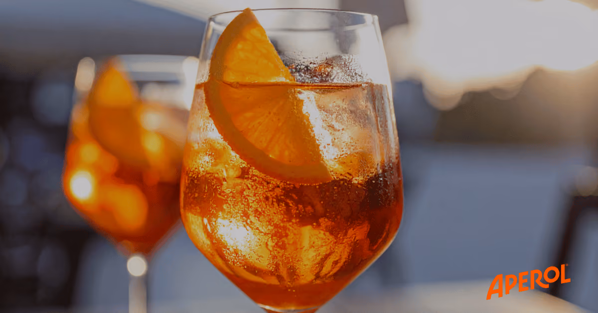 A glass of orange Aperol Spritz with ice and an orange slice, with another glass blurred in the background.