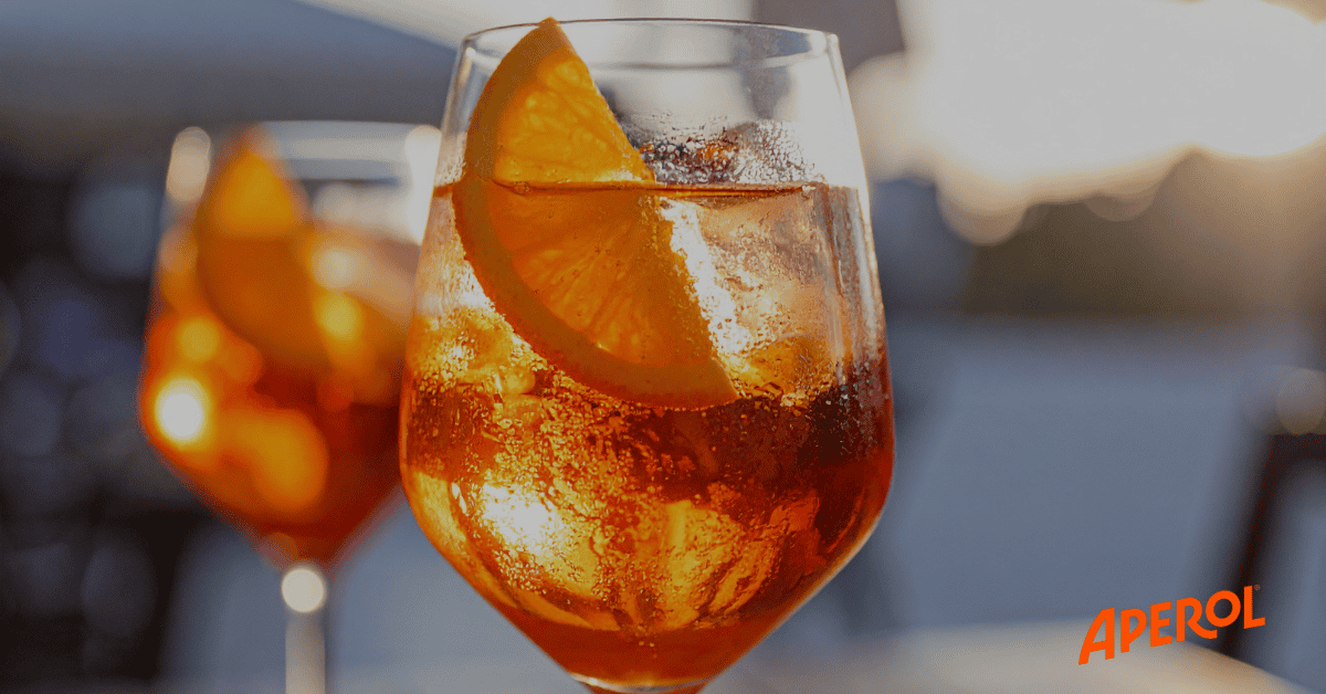 A glass of orange Aperol Spritz with ice and an orange slice, with another glass blurred in the background.