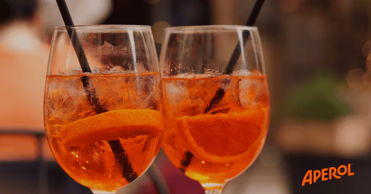 Two glasses of orange Aperol spritz with ice and orange slices, each with a black straw. Aperol logo in corner.