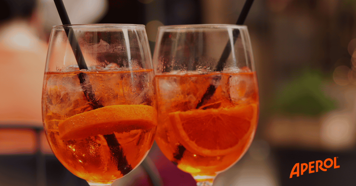 Two glasses of orange Aperol spritz with ice and orange slices, each with a black straw. Aperol logo in corner.