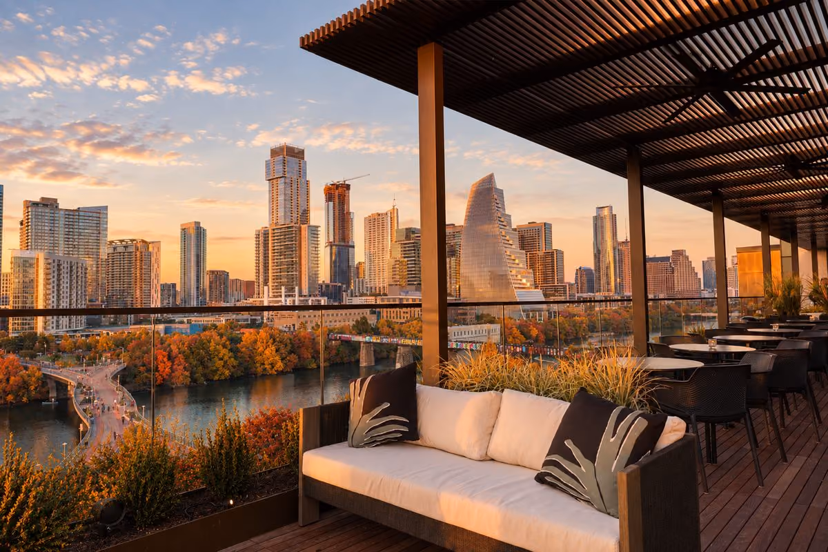 Rooftop terrace at The Loren at Lady Bird Lake with an outdoor sofa and dining tables overlooking fall foliage, the lake, and the downtown Austin skyline at sunset
