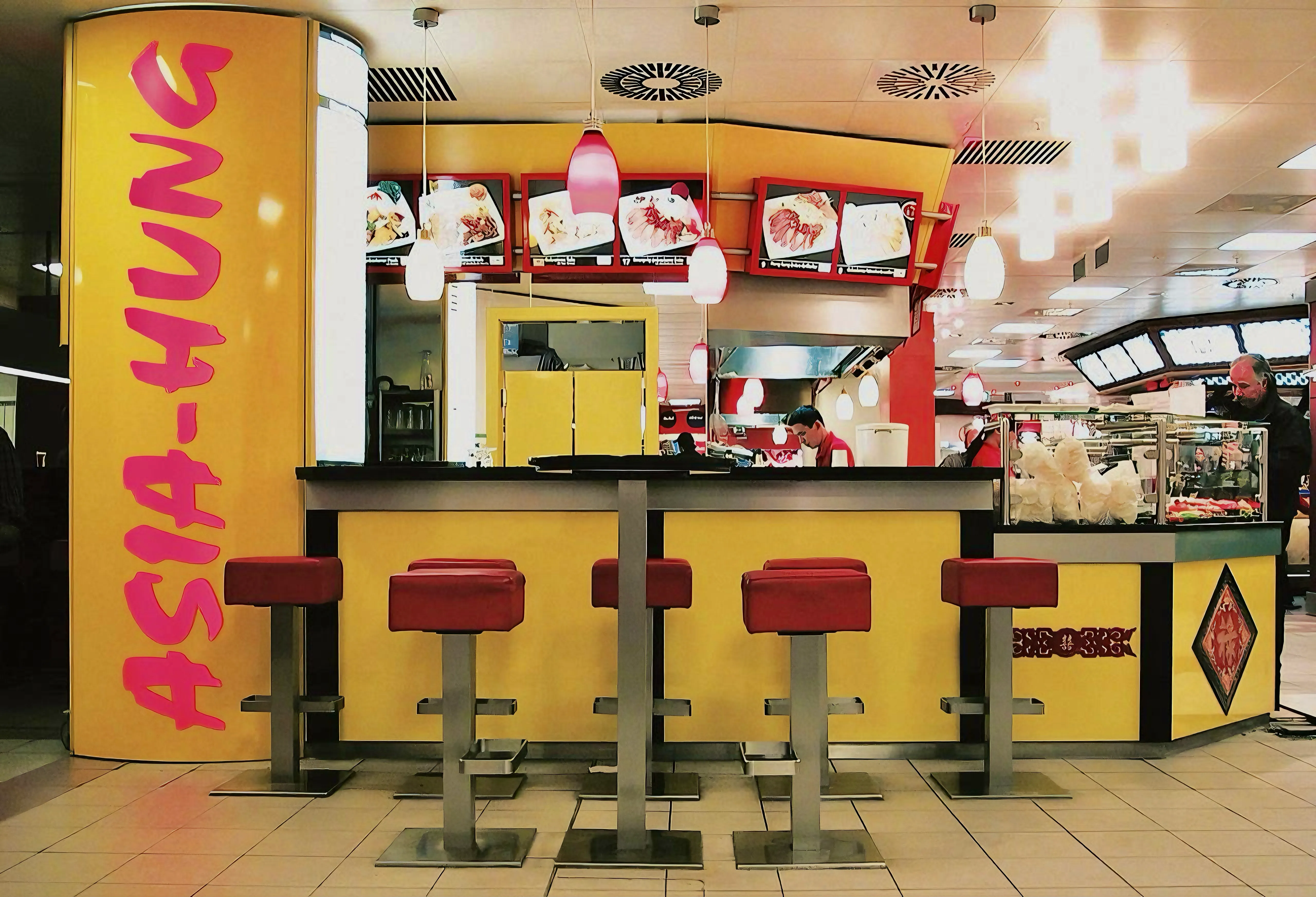 Innenansicht eines asiatischen Schnellrestaurants mit gelbem Tresen, rotem Schriftzug Asia-Hung und mehreren roten Barhockern.