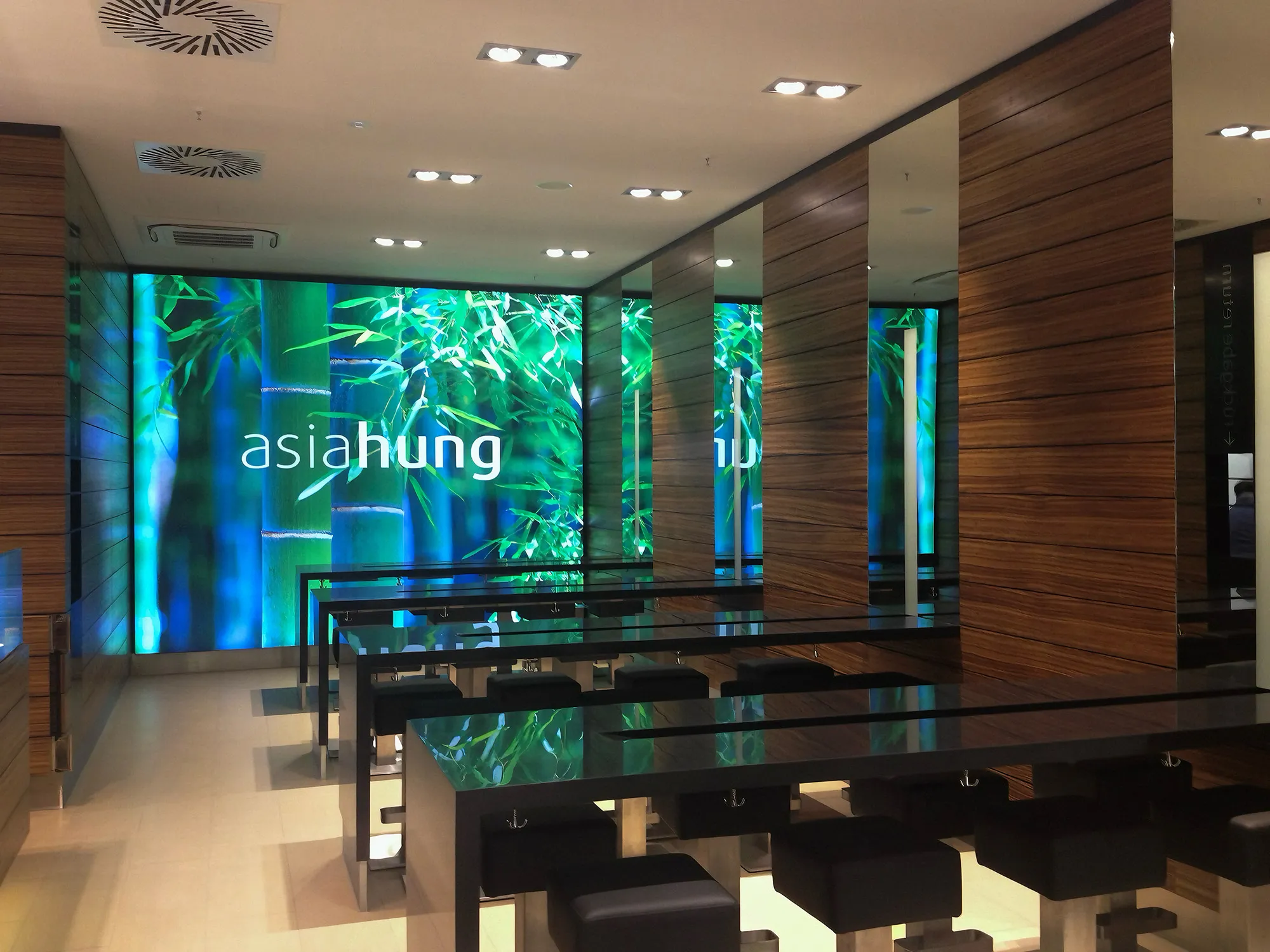 Moderne, minimalistische Caféeinrichtung mit schwarzen Tischen und Hockern vor einer leuchtenden Wand mit Bambus und dem Schriftzug asia hung.