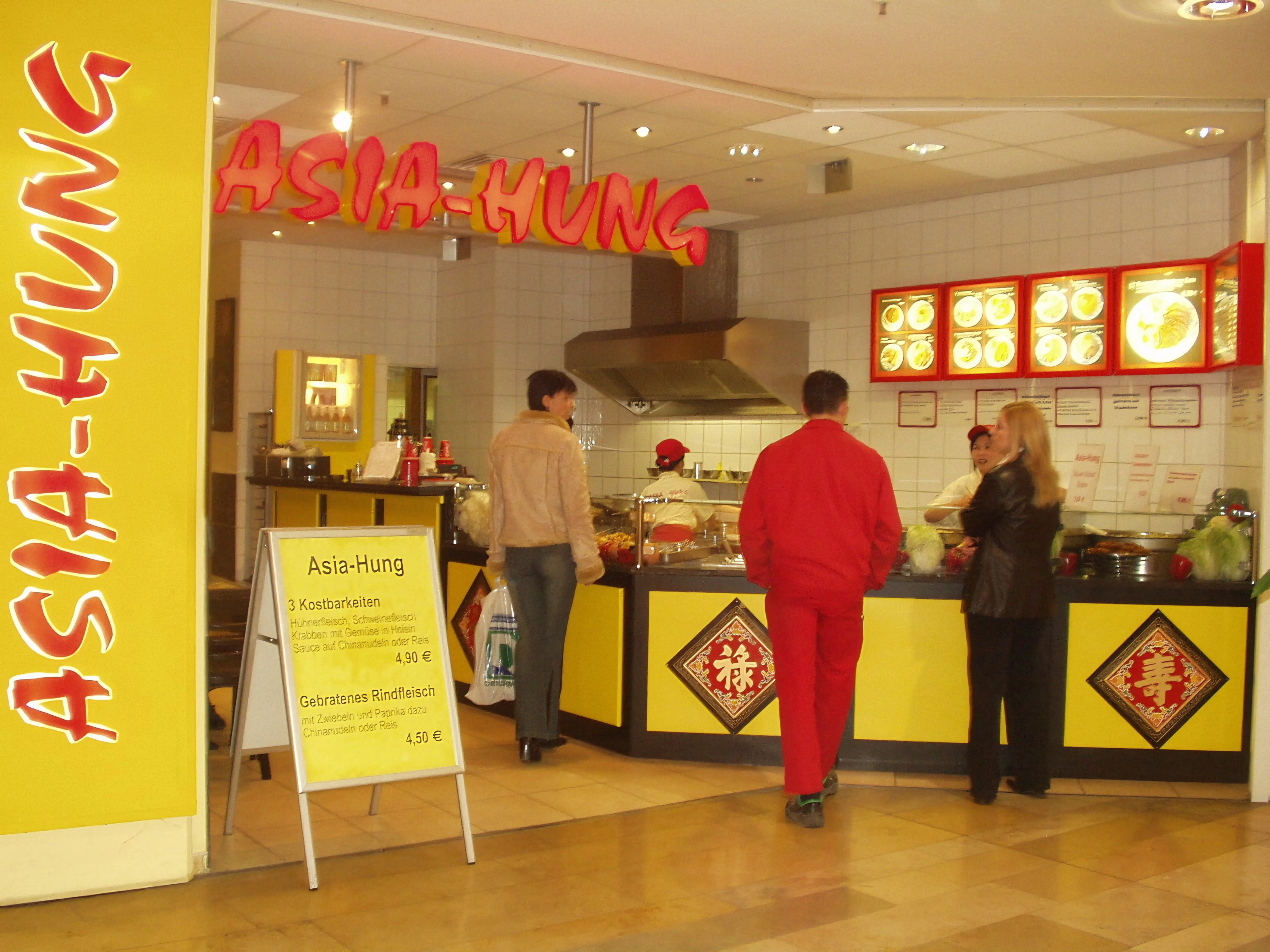 Ein kleines asiatisches Imbissrestaurant mit gelben und roten Dekorationen, drei Kunden stehen am Tresen, eine Speisekarte auf einem Ständer zeigt Gerichte und Preise.