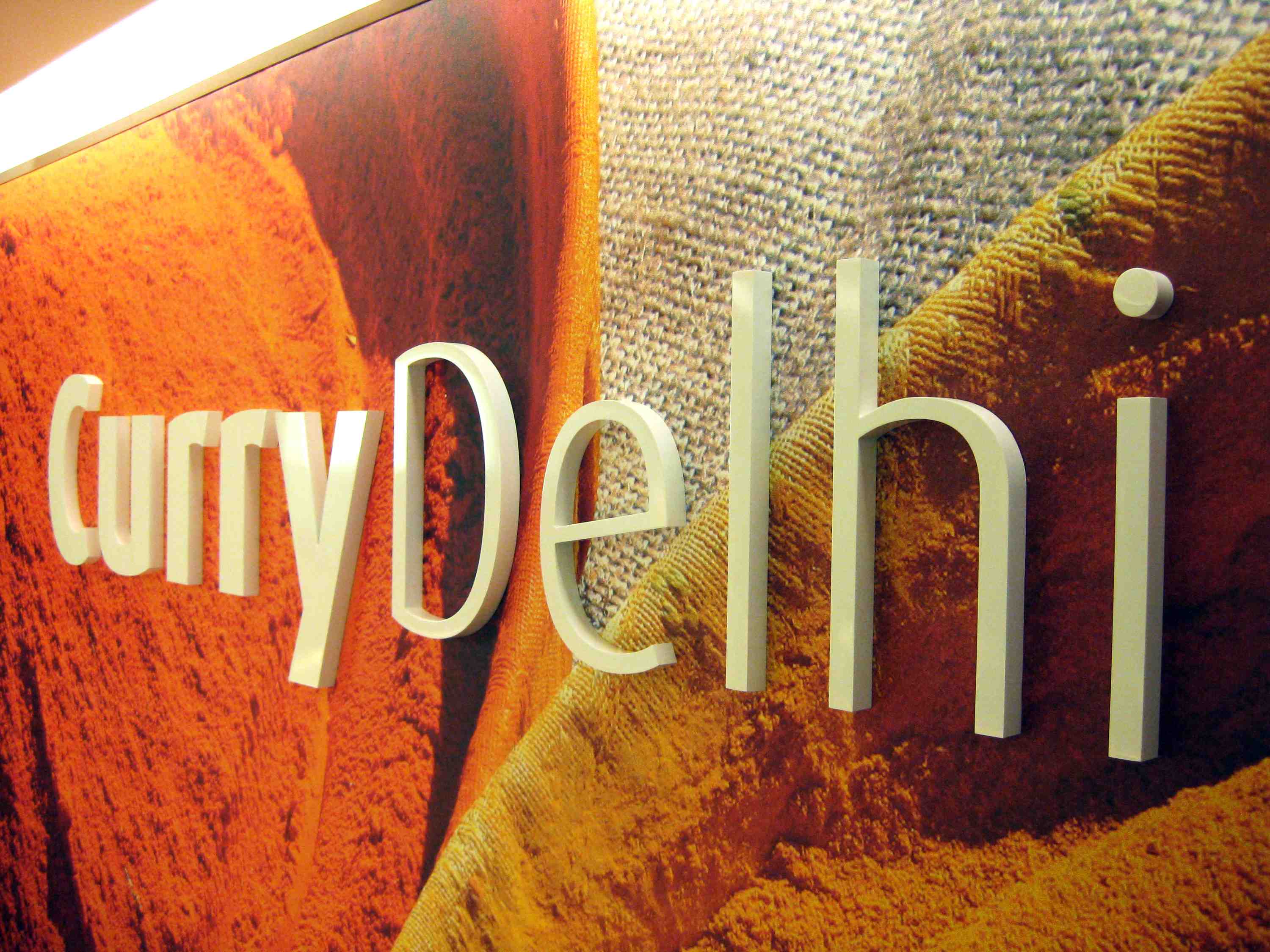 Reliefschrift "Curry Delhi" auf einer Wand mit orangenen Gewürzpulvern im Hintergrund.