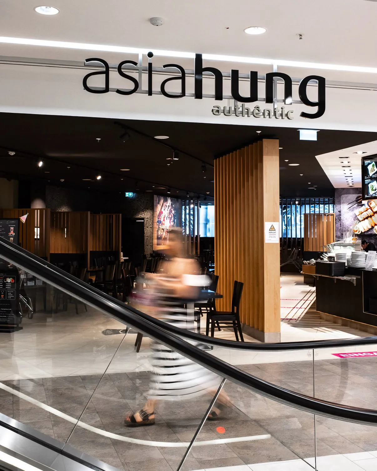 Eingang zum Asia Hung Restaurant mit verschwommener Person, die an einer Rolltreppe vorbeigeht.