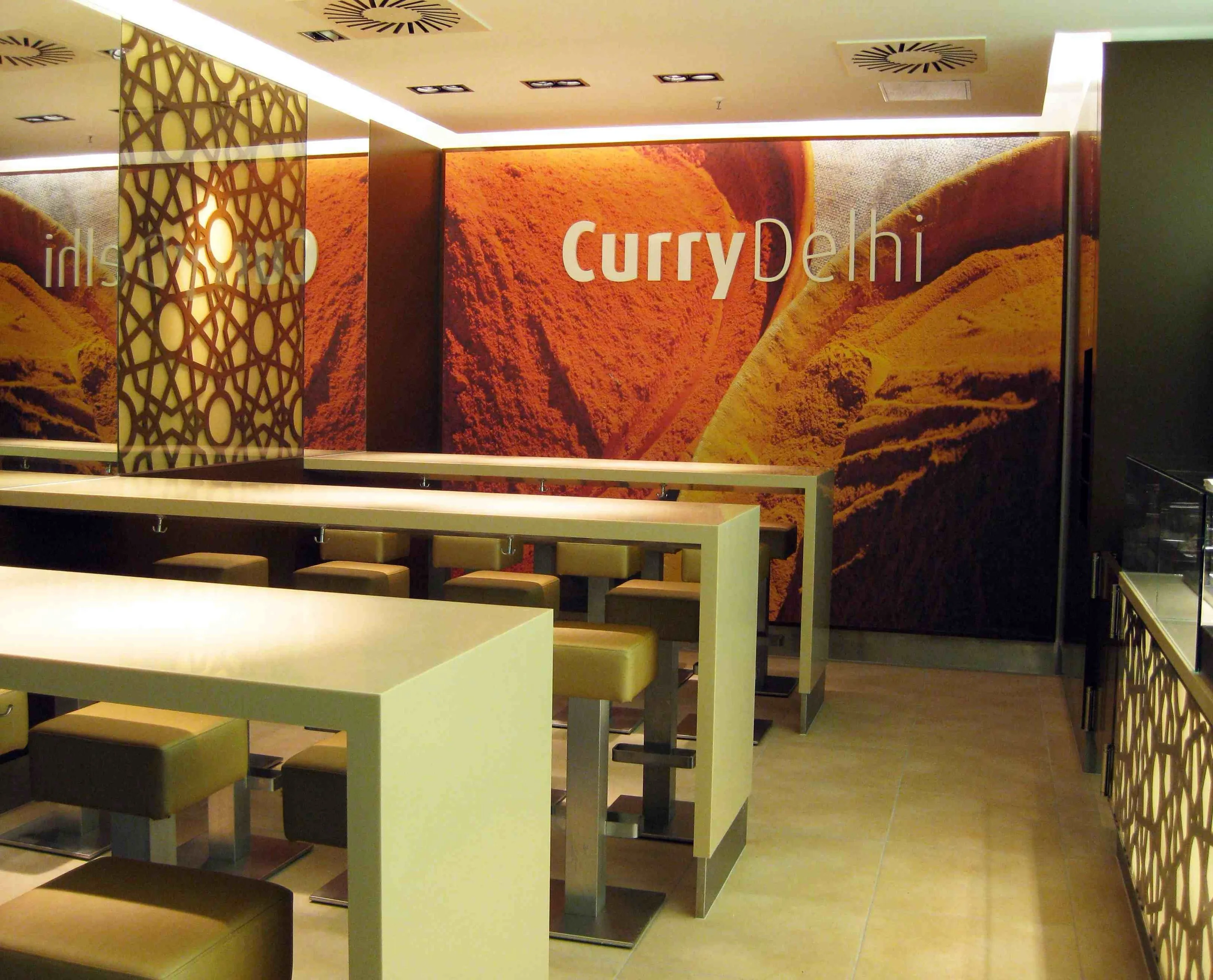 Modernes Restaurant mit hellen Tischen und Hockern vor einer Wand mit Curry-Gewürzbild und Schriftzug Curry Delhi.