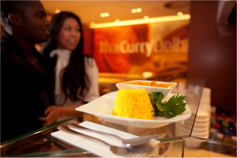 Vorne auf einem weißen Teller ist ein gelber Reis mit Koriander, dahinter steht eine Schale mit Thai-Curry, unscharf im Hintergrund zwei Personen in einem Restaurant.