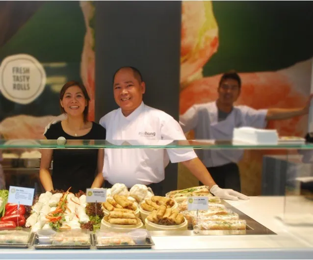 Zwei lächelnde Mitarbeiter hinter einer Glasvitrine mit frisch zubereiteten Frühlingsrollen und anderen asiatischen Snacks in einem Imbiss.